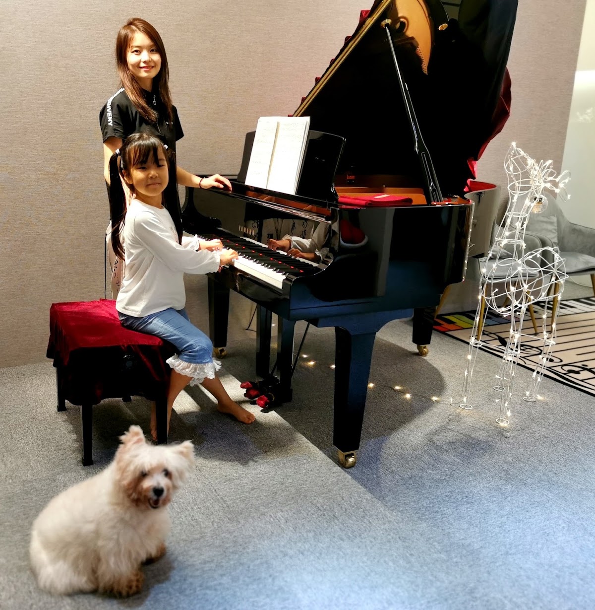 A young student playing the grand piano beside her teacher, with the studio dog nearby